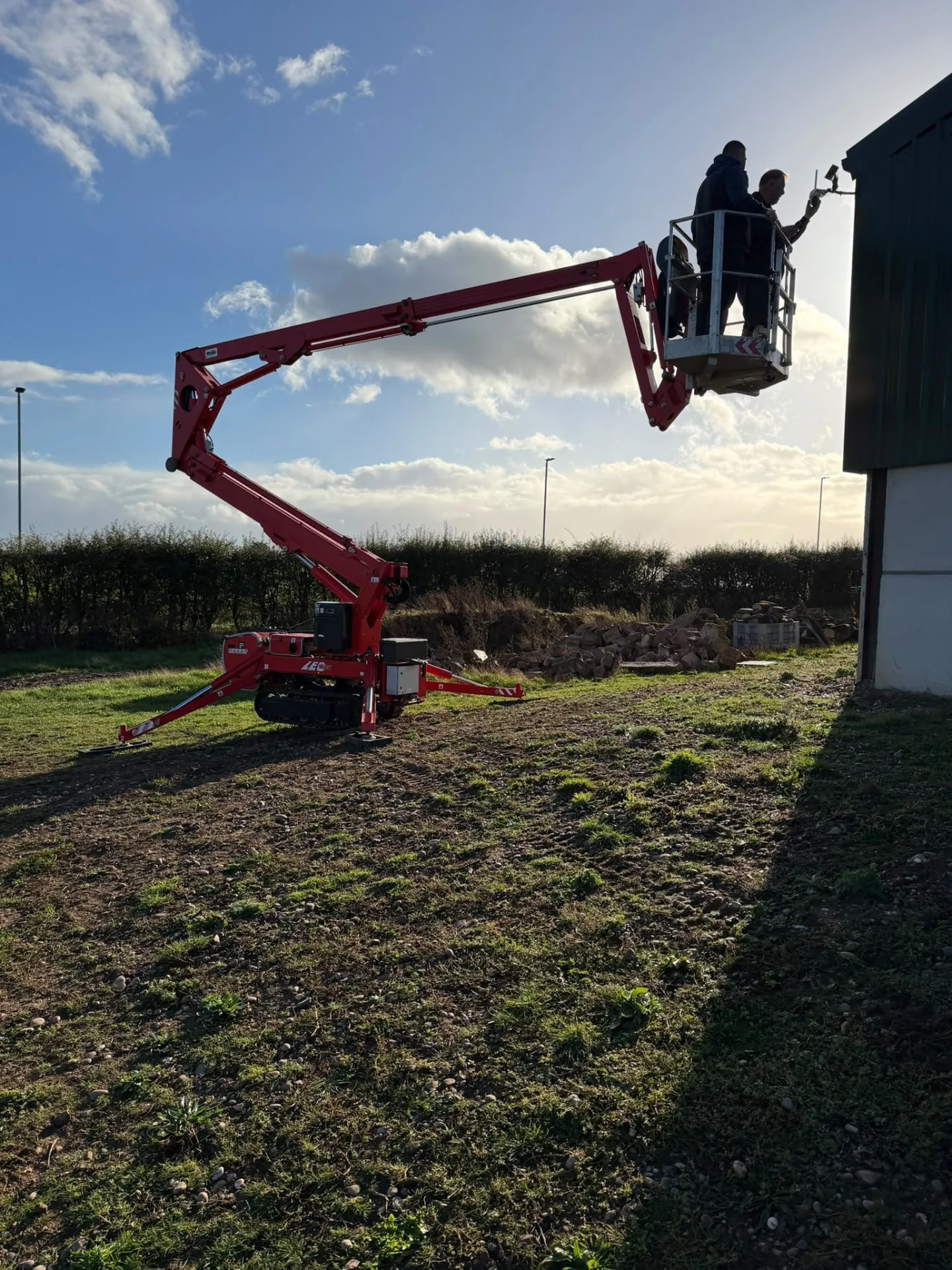Operated cherry picker hire: two people in the basket fitting equipment to the eaves of a farm building